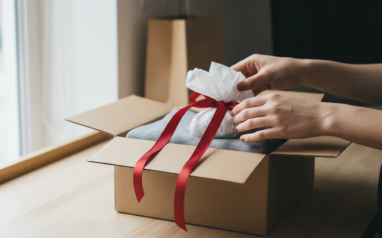 Mãos colocando um pequeno pacote com fita vermelha sobre uma peça de roupa dentro de uma caixa de envio